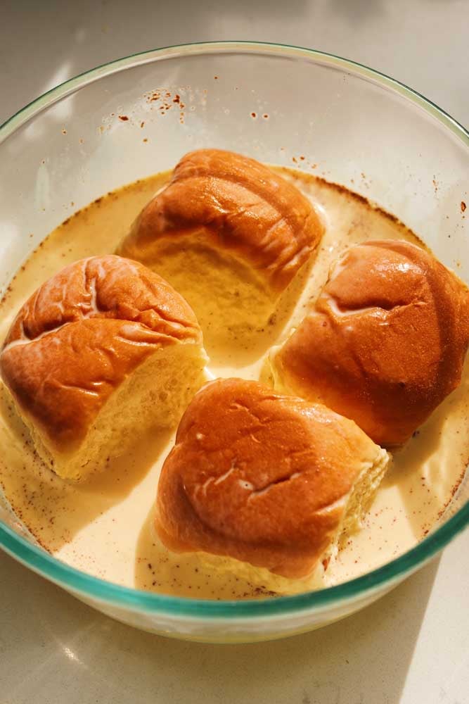 four hawaiian rolls submerged in a glass bowl filled with french toast batter.
