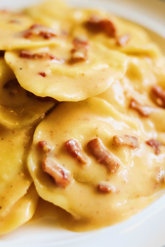 an up close view of two raviolis covered in a yellow carbonara sauce with specs of red bacon. 