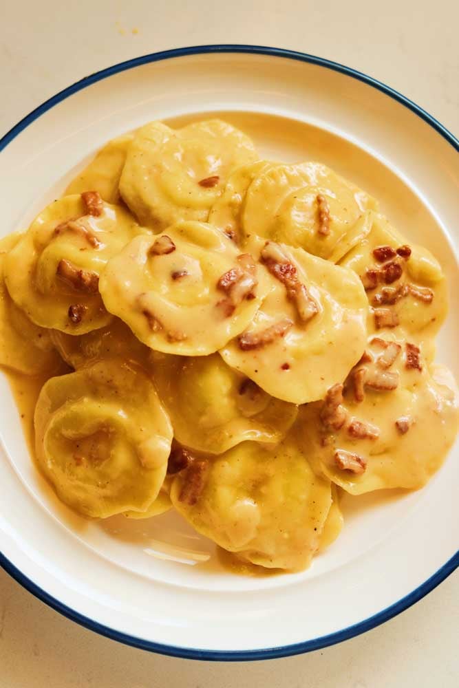 a blue and white plate filled with creamy yellow carbonara covered cheese ravioli. 