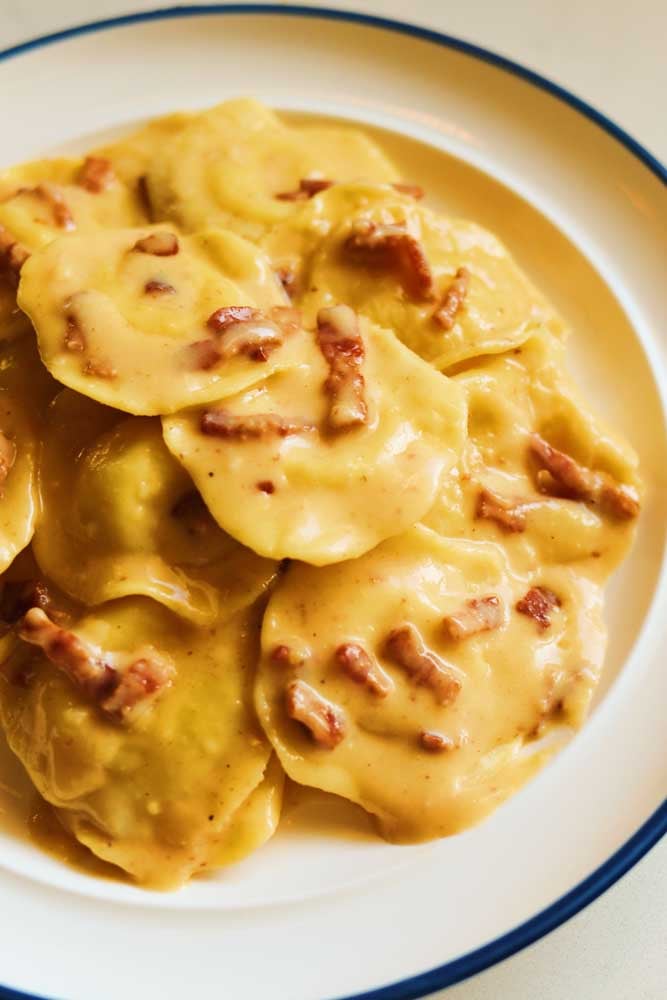 the right side of a white plate topped with ravioli covered in a yellow carbonara sauce with specks of red bacon throughout. 