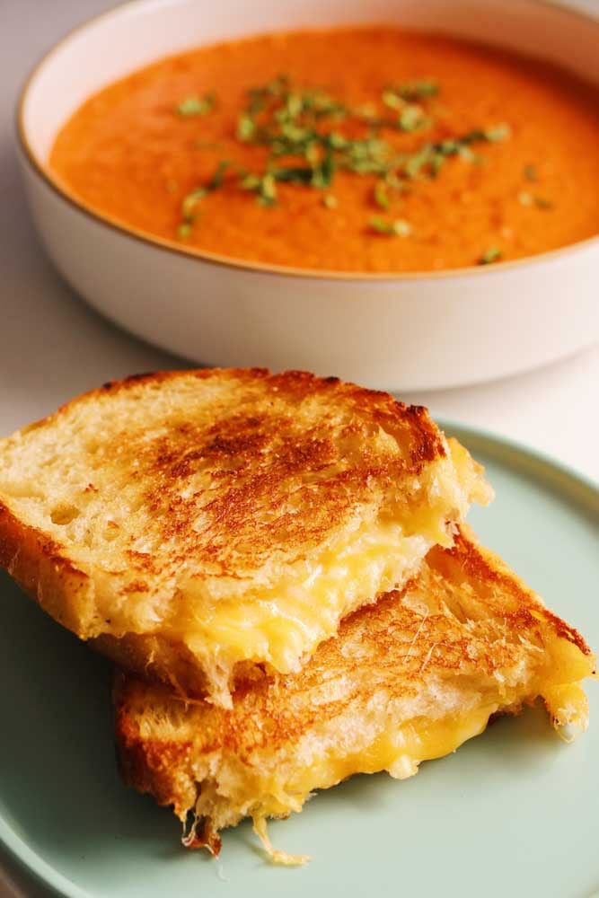 a blue plate topped with two grilled cheese halves with a bowl of tomato soup in the back. 