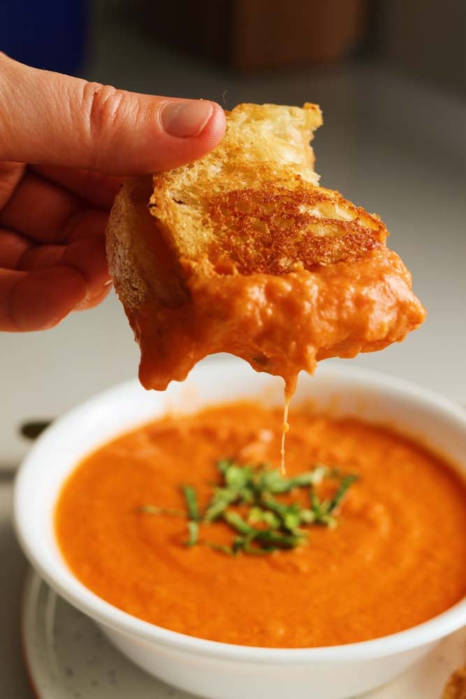 a hand holding a piece of grilled cheese dunking it into a bowl of tomato soup with basil on top. 