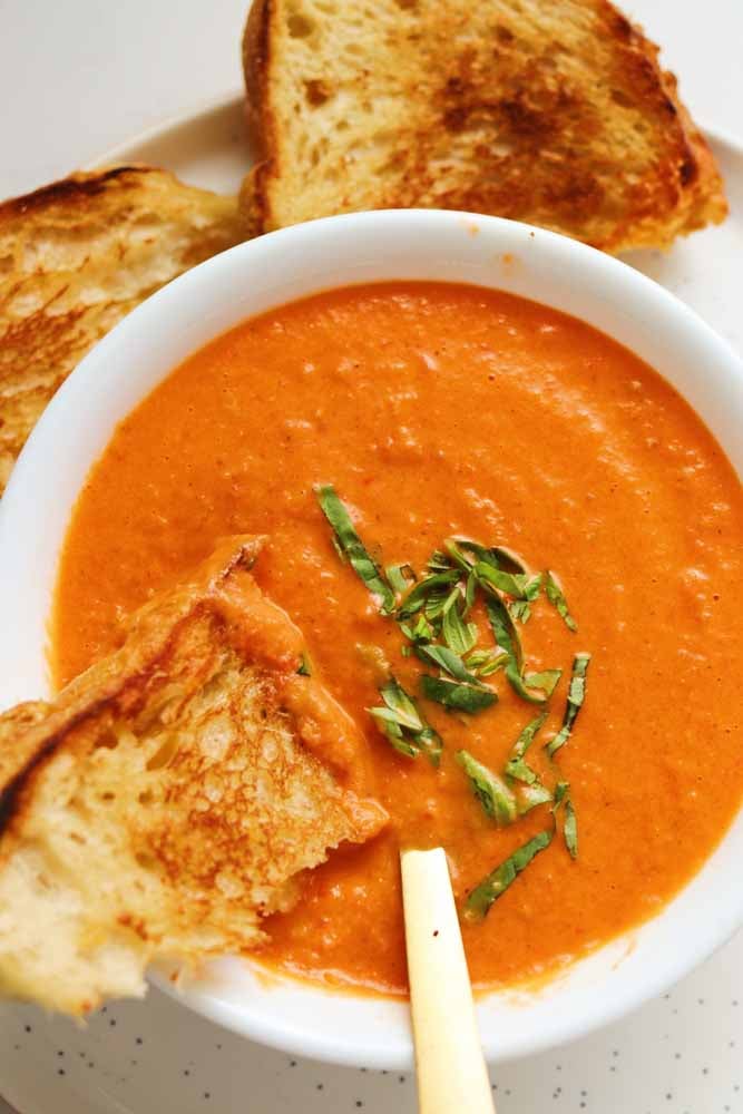 a white bowl full of tomato soup with basil on top with a grilled cheese sitting behind it. 