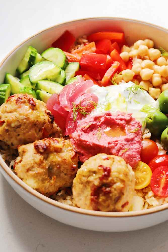 a close up view of the left side of a white bowl filled with mediterranean chicken meatballs, feta rice, fresh veggies and colorful dips.