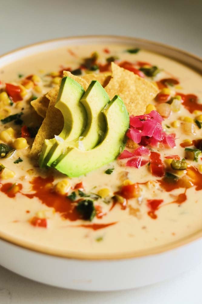 a white bowl filled with warm soup made of corn, peppers and cheese with avocado, pink pickled onions and chips on top.