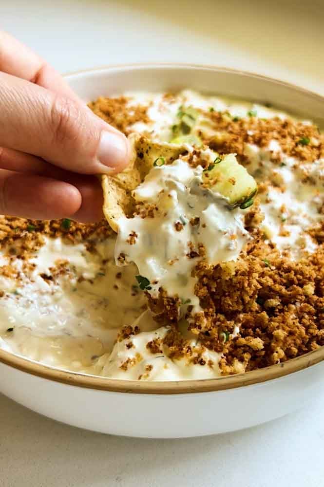 a hand scooping a tortilla chip into a bowl of fried pickle dip. 