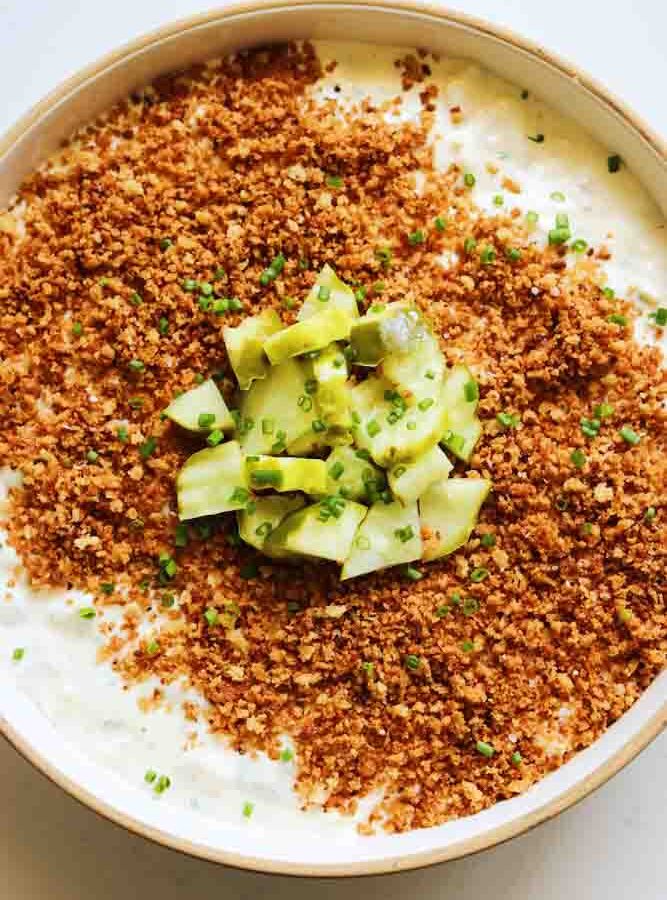 a white bowl of creamy white pickle dip with crunchy brown bread crumbs and chopped pickles on top.