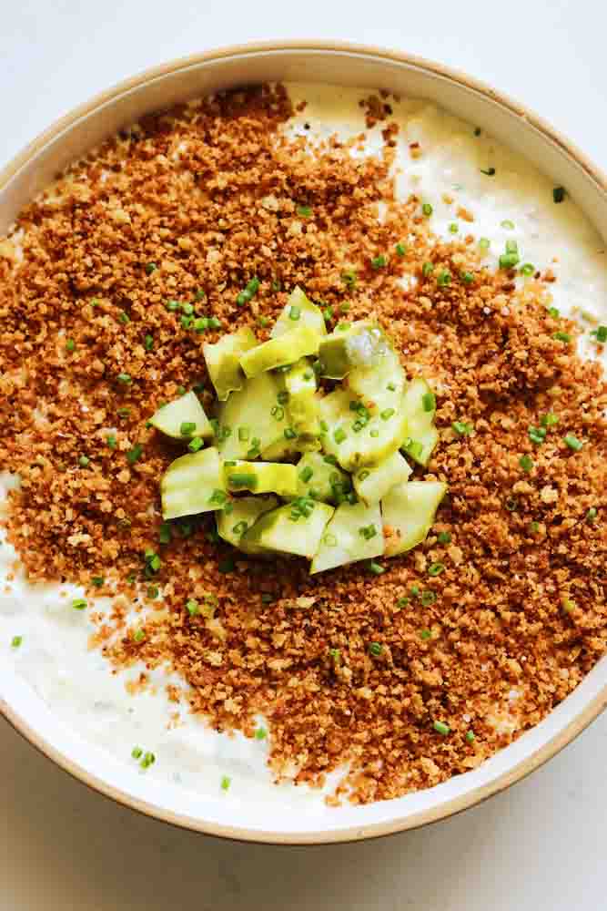 a white bowl of creamy white pickle dip with crunchy brown bread crumbs and chopped pickles on top. 