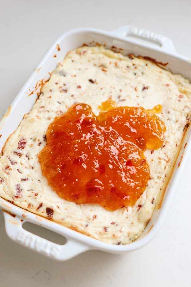 A baking dish full of cheese dip with apricot pepper jelly smeared on top about to be smeared on. 