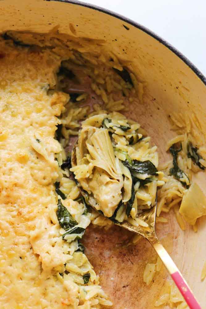 an up close view of a bowl of spinach artichoke orzo with a scoop taken out of it. 