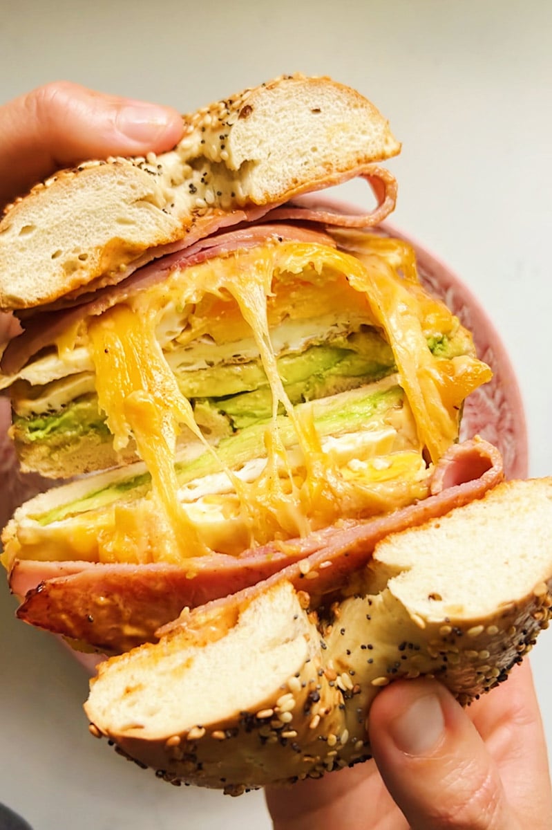 a ham egg and cheese bagel being split in half with a cheese pull happening in the middle. 
