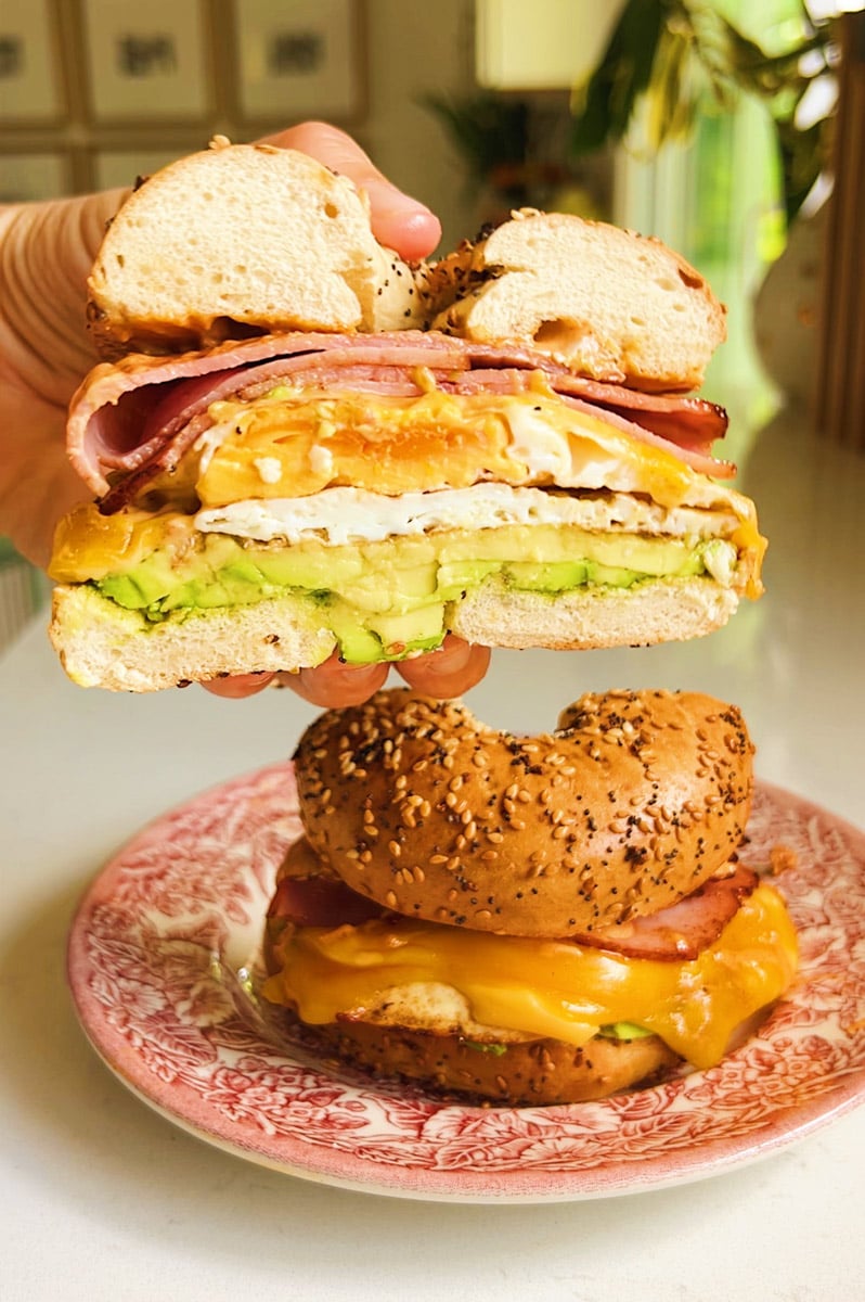 a hand holding half of a bagel sandwich over a plate with the other half. 