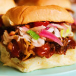 an up close view of a pulled pork slide with coleslaw, sauce and pickled red onions on hawaiian rolls.