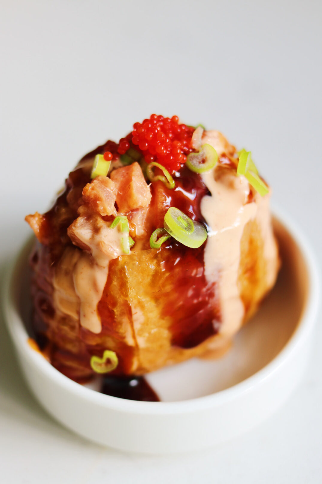 a singular piece of spicy tuna inari in a tiny bowl topped with fish roe and scallions, spicy mayo, and unagi eel sauce.