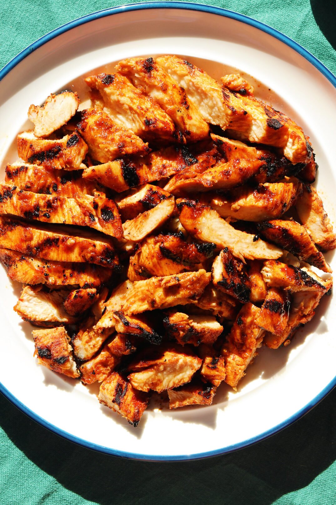 a white plate full of sliced harissa chicken with a sweet honey glaze on a teal tablecloth. 