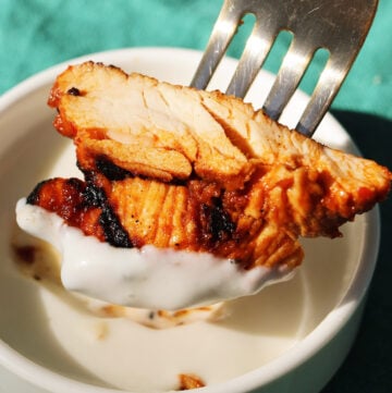 a fork holding a slice of harissa chicken dunking it into white garlic sauce.