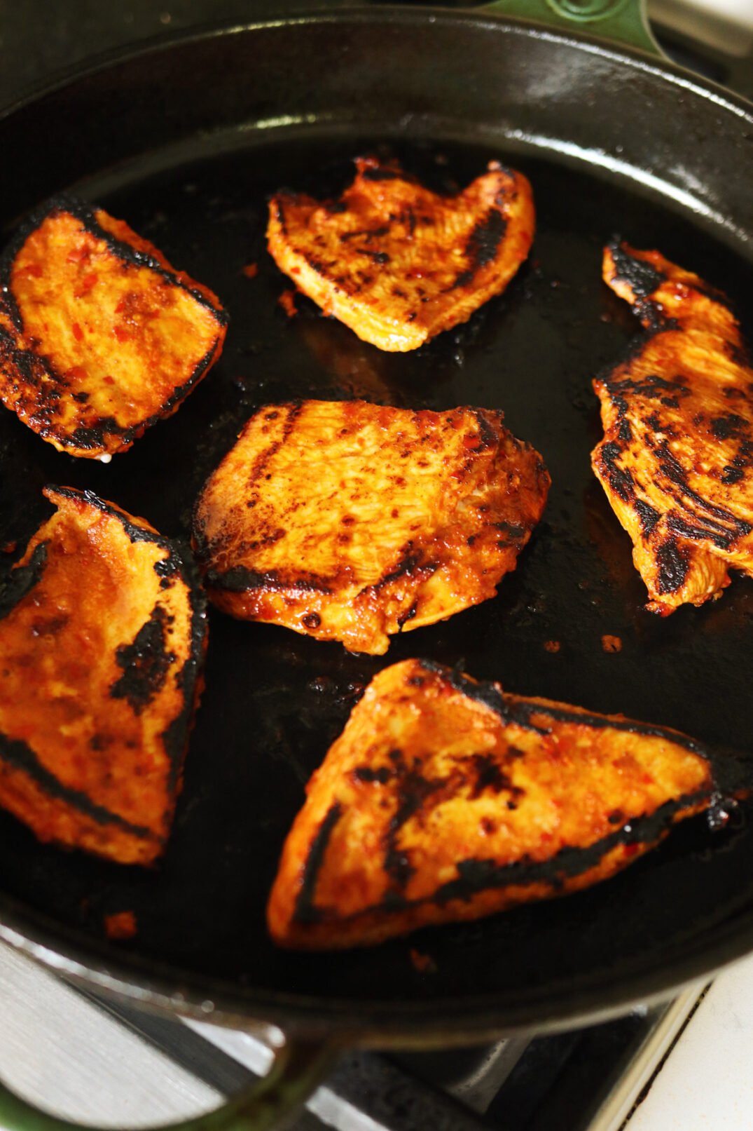 pieces of seared harissa chicken cooking in a large green cast iron skillet. 
