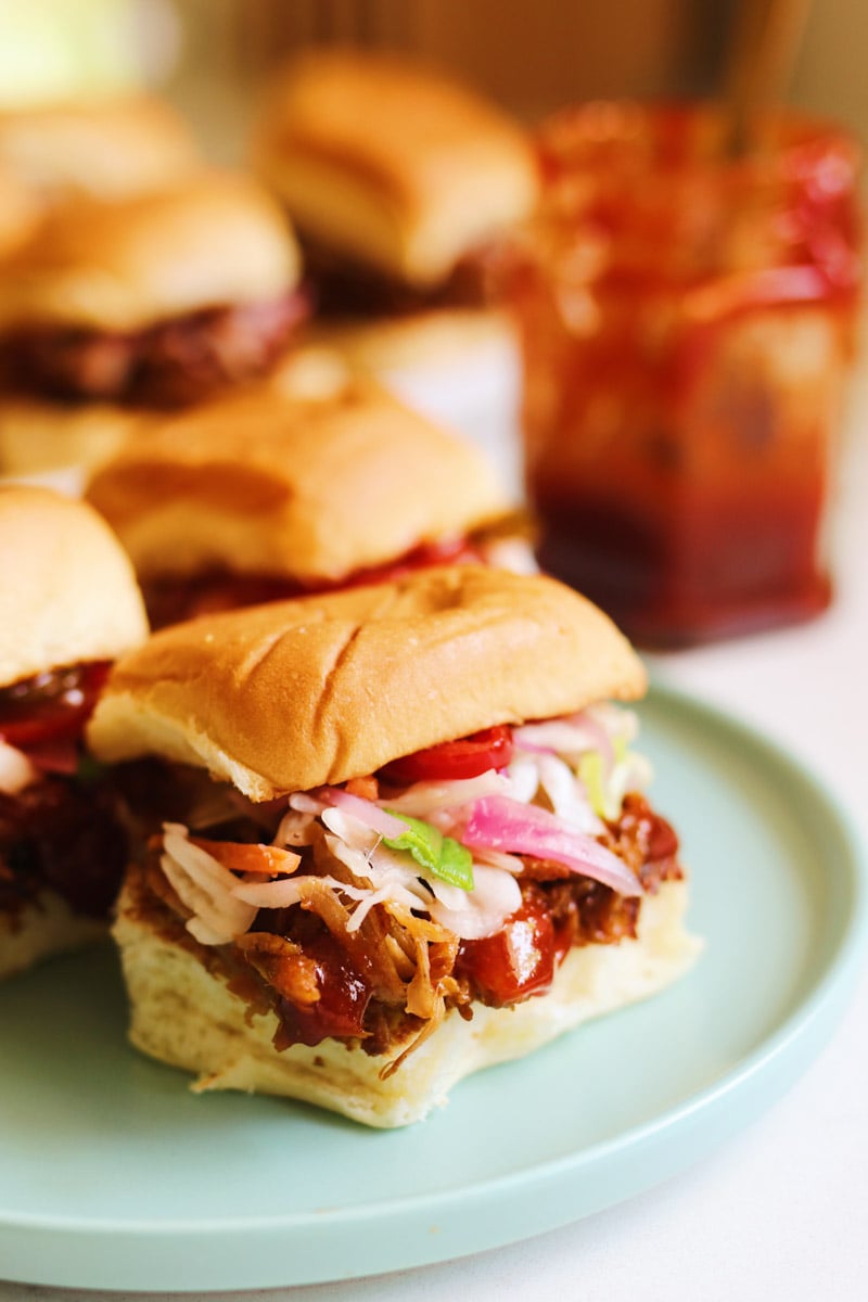 a small blue plate topped with a couple colorful pulled pork sliders with a jar of bbq sauce in the background.