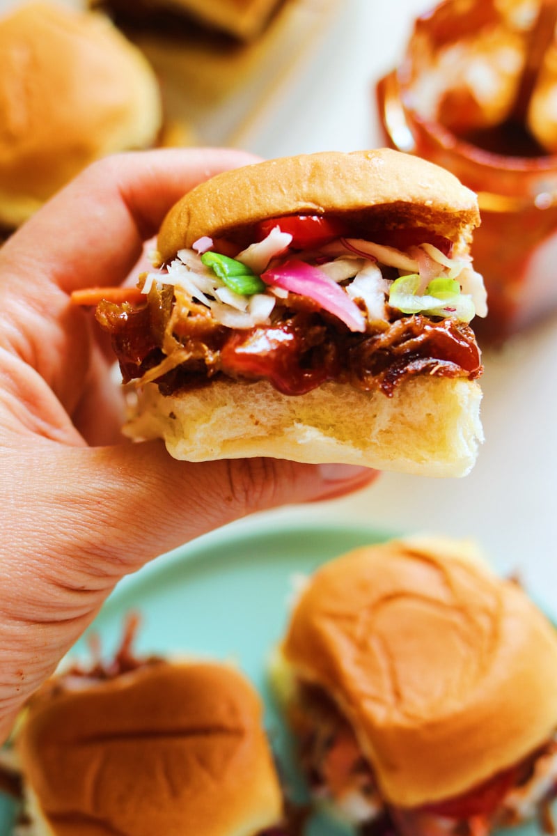 a hand holding a little pork sandwich over a plate of food.