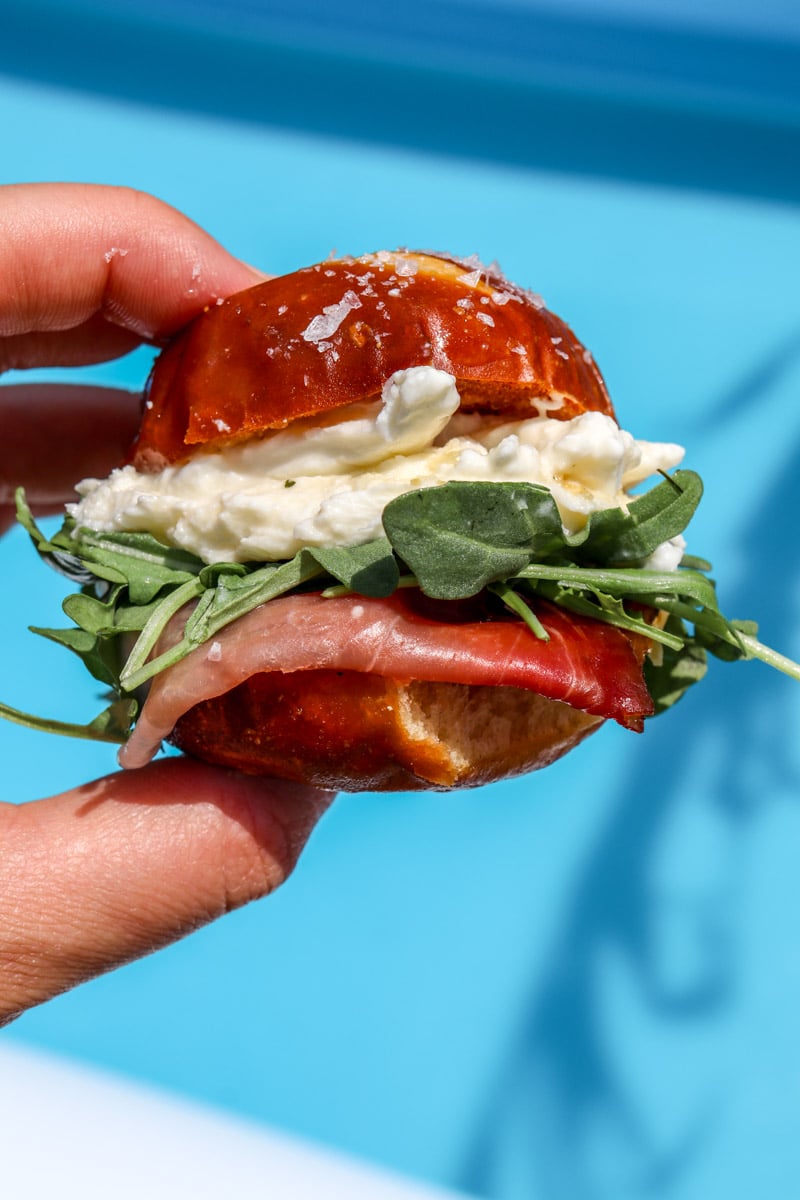 a hand holding some burrata and prosciutto sliders over a swimming pool.