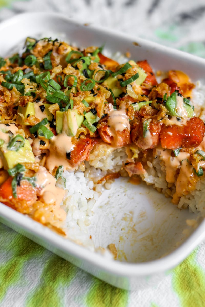a casserole dish with teriyaki salmon sushi and colorful toppings with a scoop taken out of it.