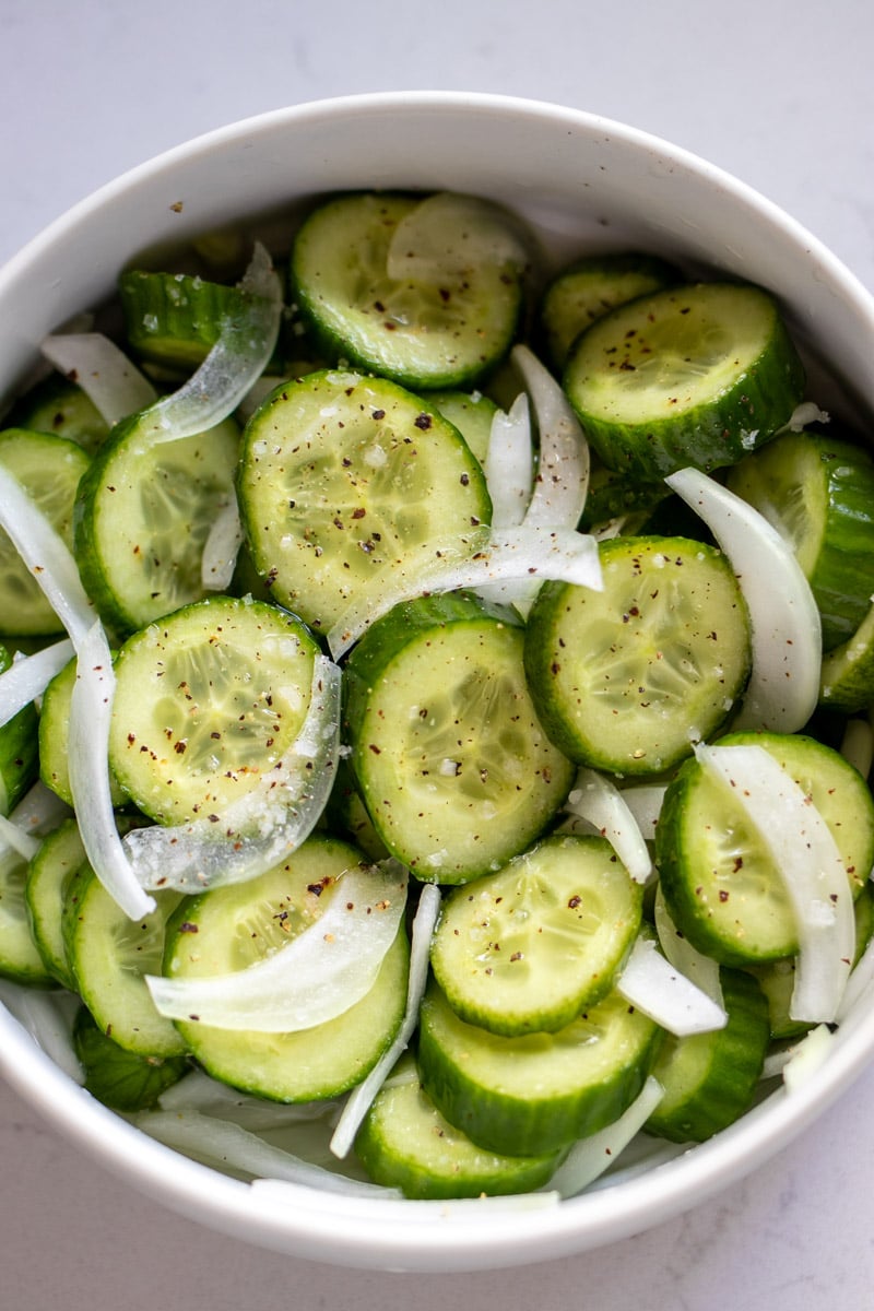 an overhead view of a white bowl of cucumber salad with thinly sliced onions on a white countertop. 