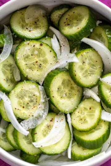 cucumber salad with sliced onions, salt, pepper in a white bowl with a pink background.