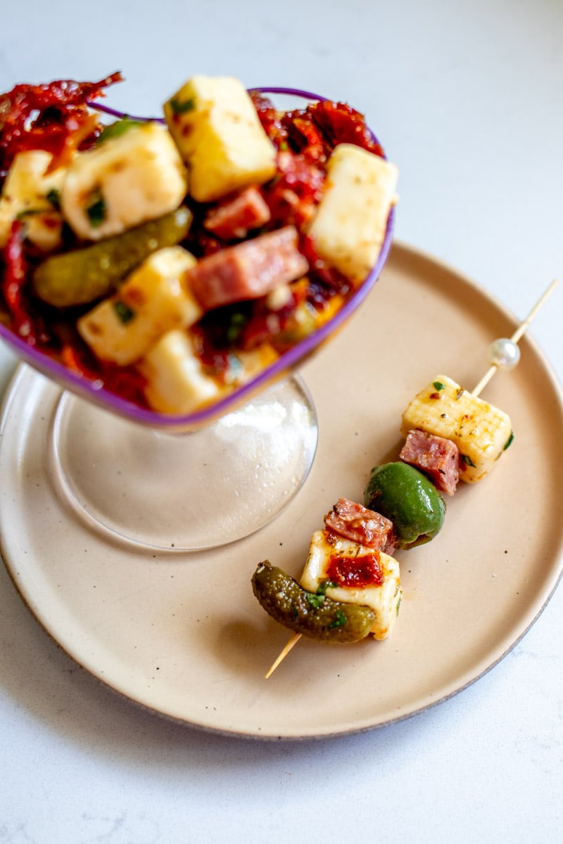 a heart shaped glass full of marinated cheese, salami and pickles with a skewer below it.