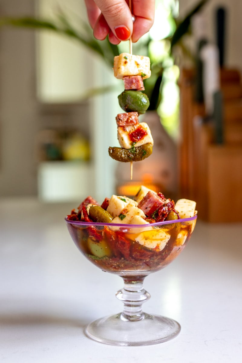 a fancy cup full of marinated cheese, olives and salami with a skewer over the top. 