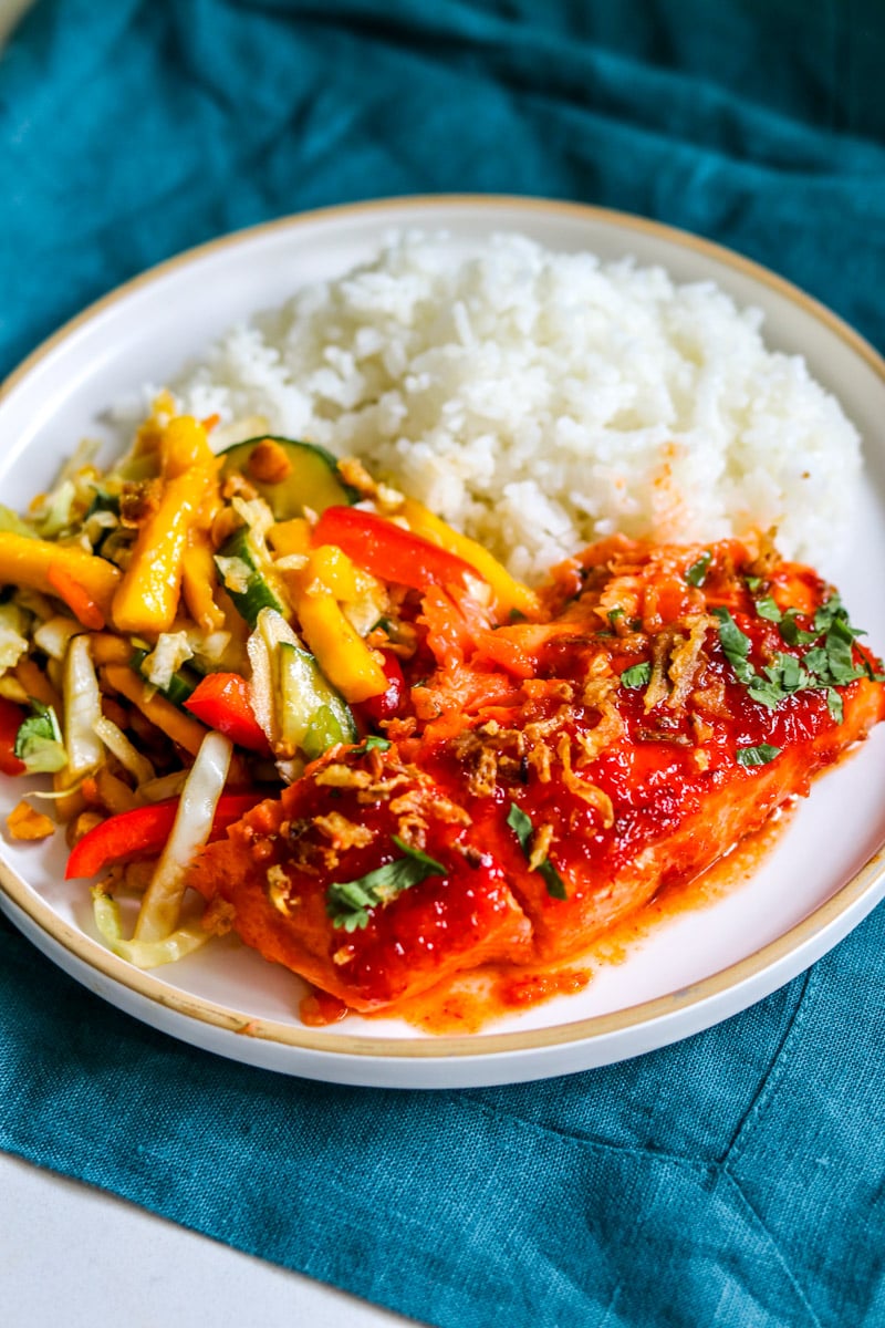 a white plate filled with a cut of spicy salmon, white rice, and mango salad. 