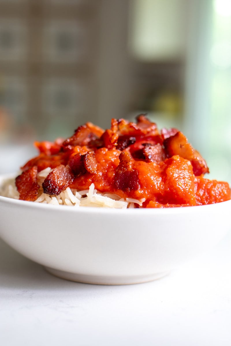 a white bowl filled with tomato gravy and bacon over rice. 