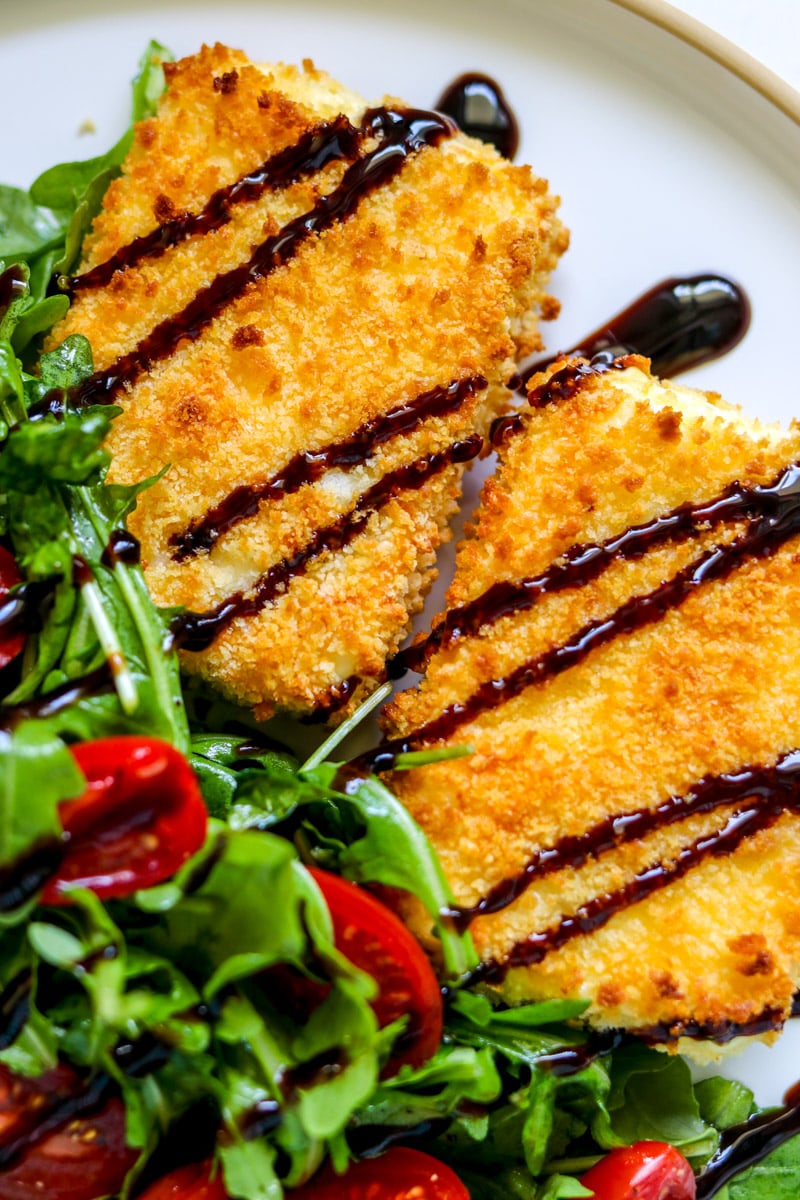 two pieces of feta milanese on a white plate with a drizzle of balsamic vinegar.