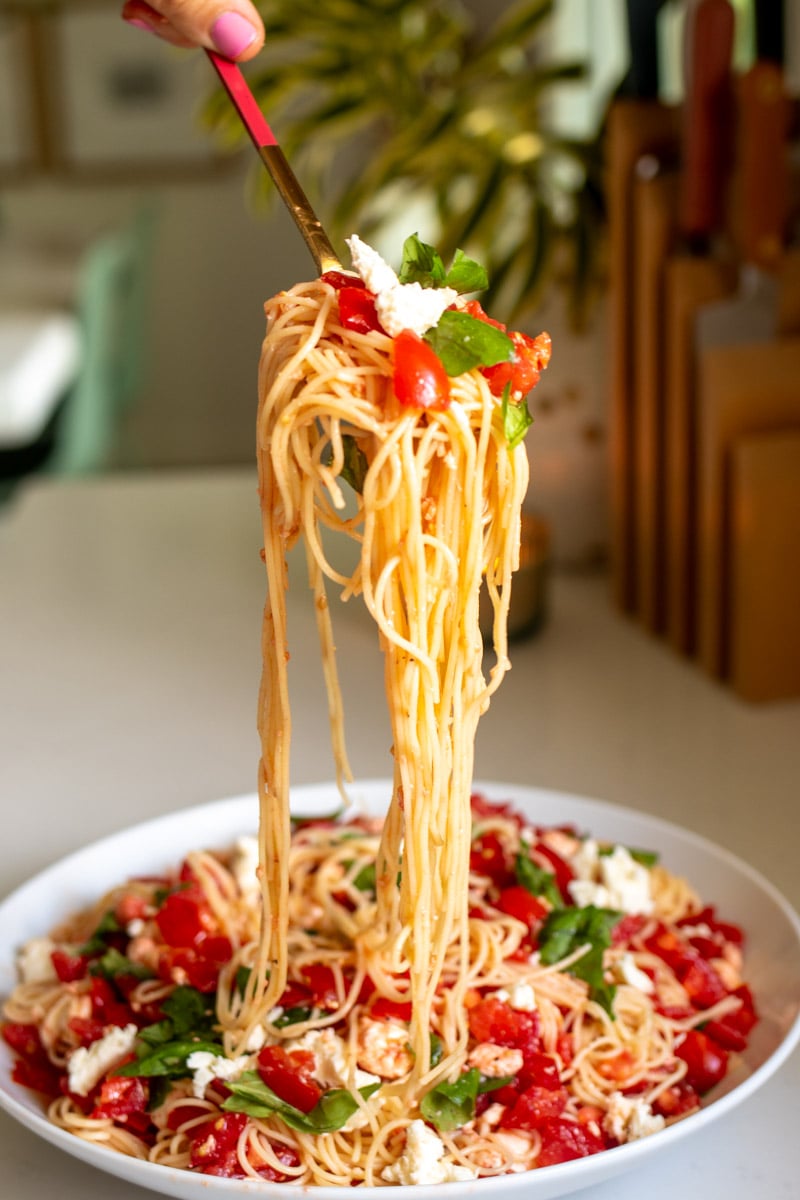 a fork scooping some bruschetta spaghetti salad over a white bowl of pasta.