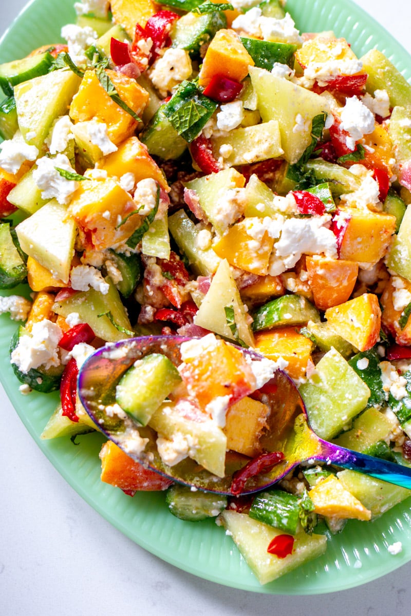 a green serving bowl filled with a colorful peach cucumber melon salad with feta. 