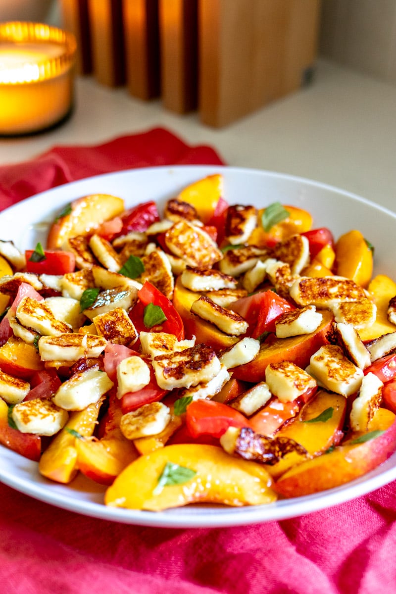 a peach tomato salad with chunks of fried halloumi on a pink tablecloth with a candle burning in the background.