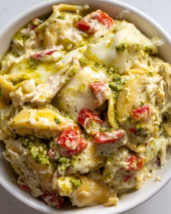 Slow Cooker Tortellini Casserole wit specks of roasted red peppers and pesto in a white bowl.