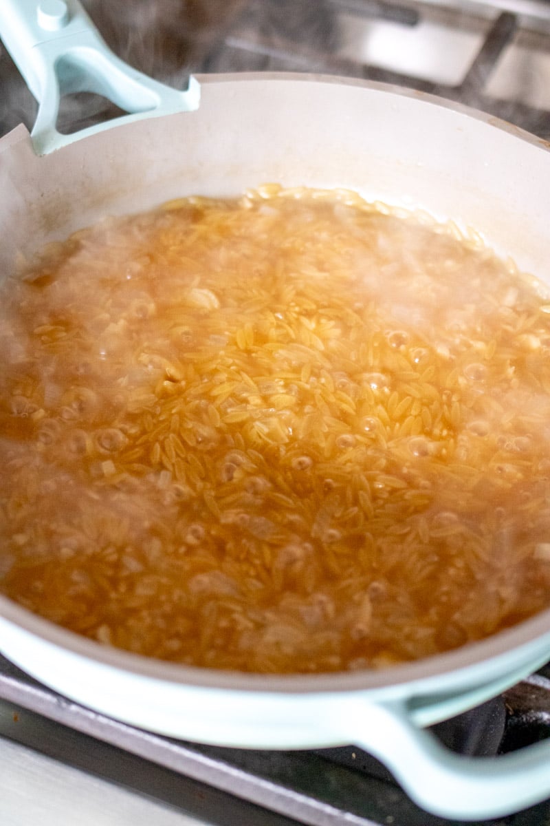 uncooked orzo with sauteed onions and garlic and broth in a blue pan.