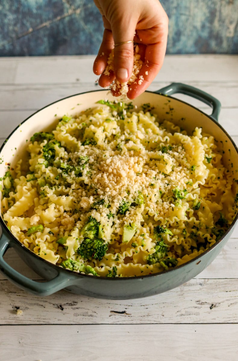 a hand sprinkling parmesan cheese on top of pasta cooked with broccoli and garlic.