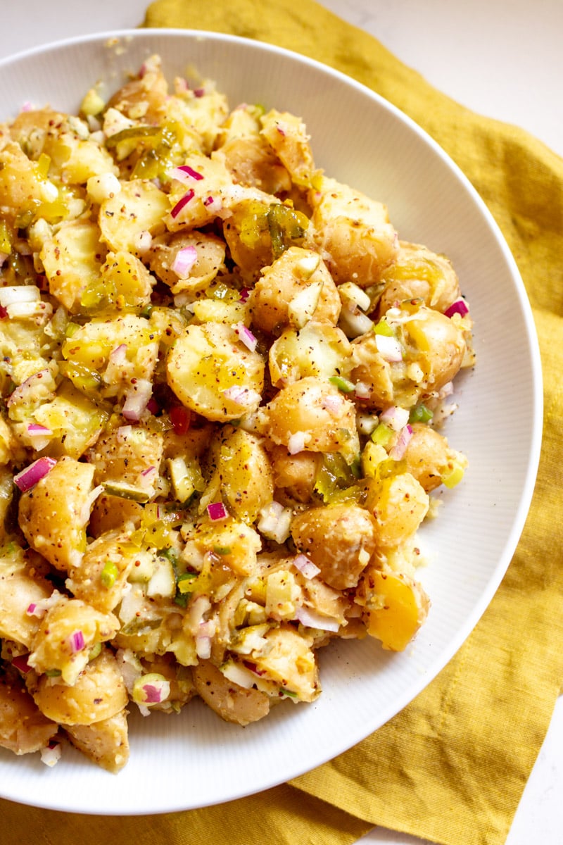 the right side of a white bowl filled with yellow potato salad with flecks of dill pickle and red onion.