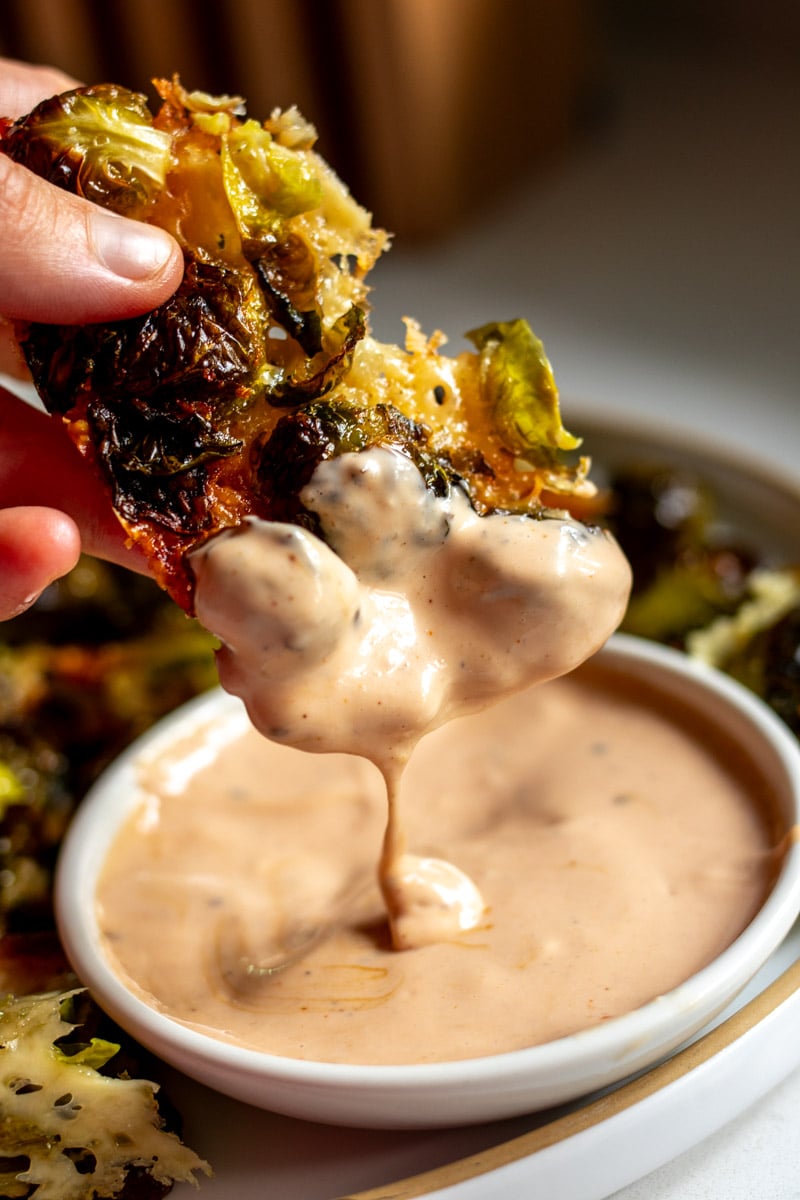 a hand dunking some parmesan crusted brussels sprouts into a thing of canes sauce.