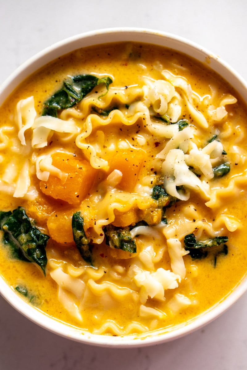 an overhead view of a white bowl filled with orange butternut squash soup with little noodles, cheese and pieces of kale popping out.