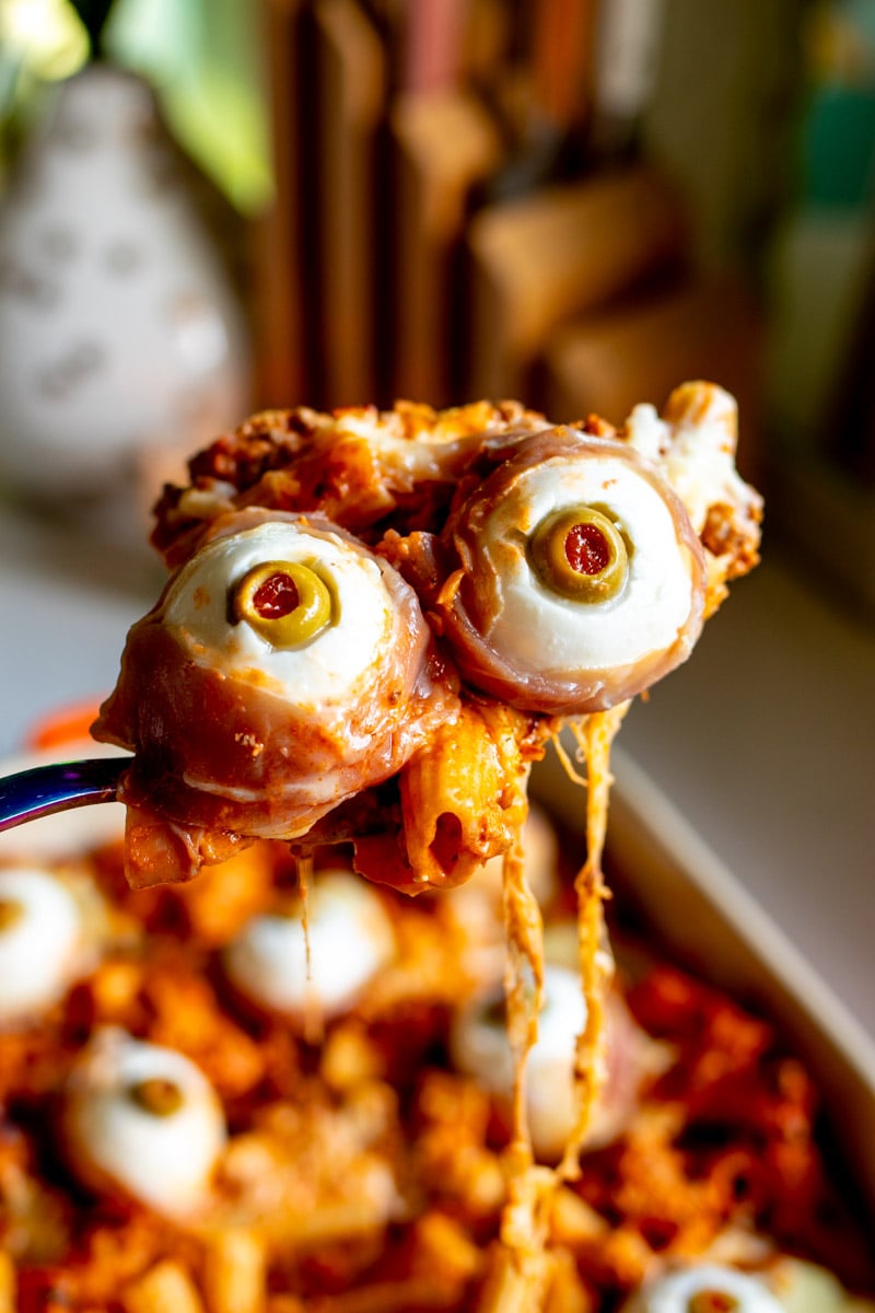 a spoon holding two mozzarella eyeballs with the baked ziti in the background.