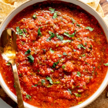 a white bowl full of red hot salsa recipe with a golden spoon and tortilla chips around it.