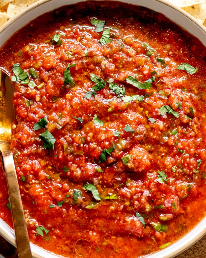 a white bowl full of red hot salsa recipe with a golden spoon and tortilla chips around it.