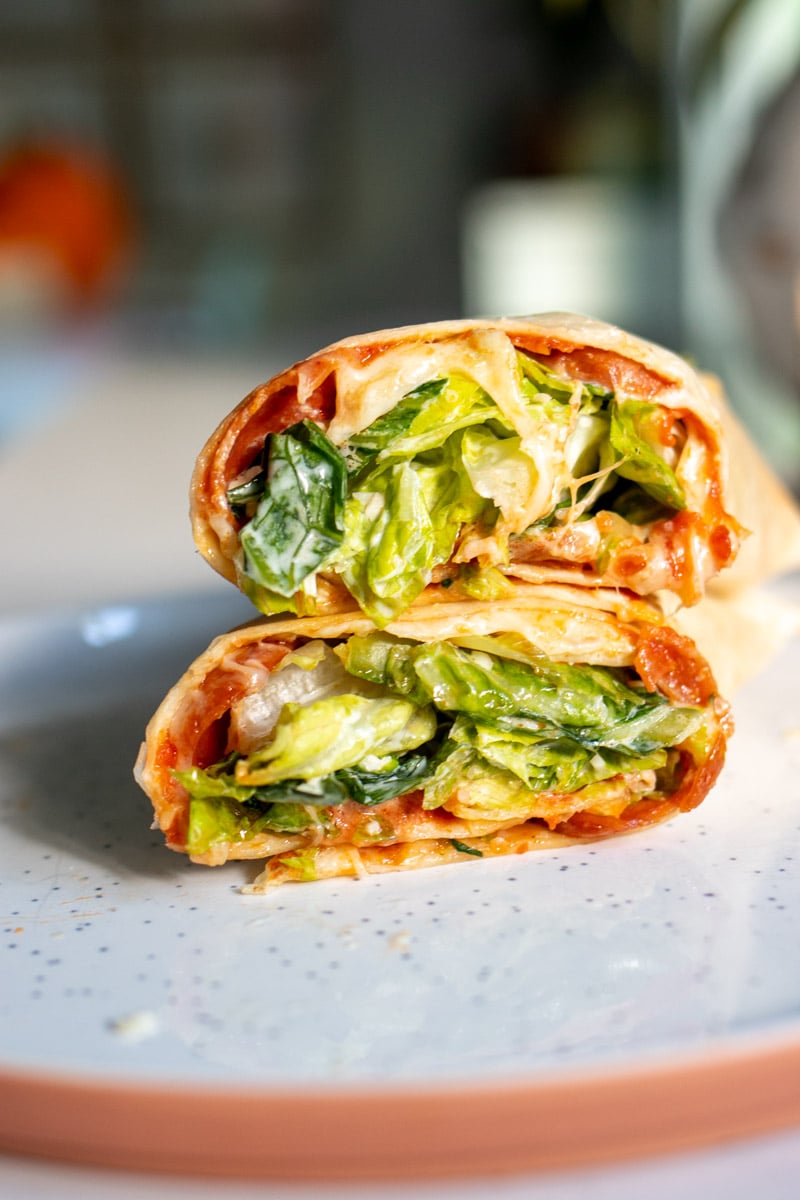 a pepperoni pizza caesar salad wrap cut in half on a white plate. 