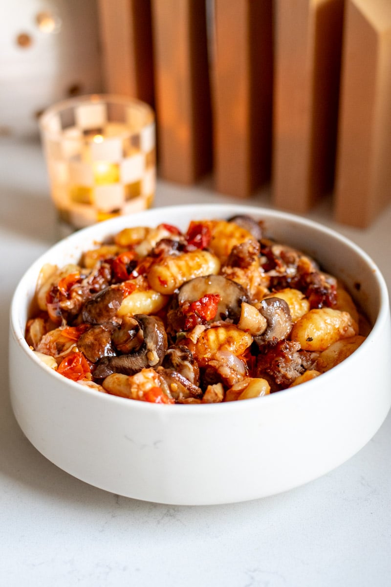 a white bowl filled with sausage, veggies and gnocchi with a candle in the background.