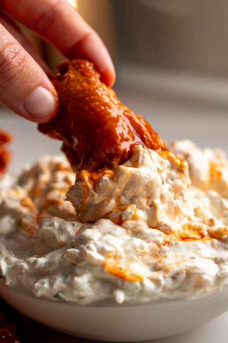 a hand diping a buffalo chicken wing into caramelized onion dip. 