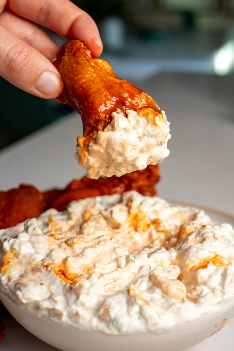 a hand dipping a wing in a white bowl of caramelized onion dip with blue cheese.