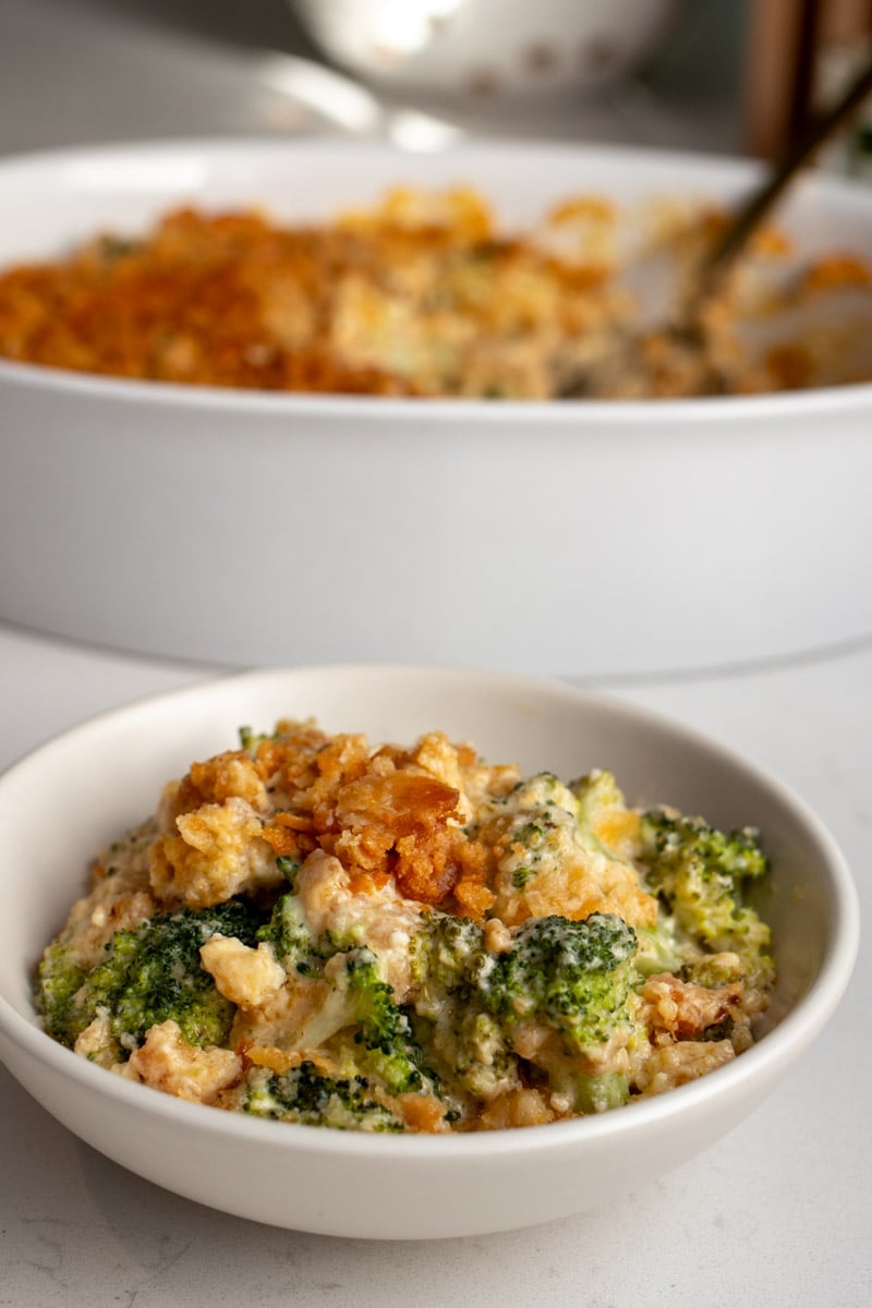 a white bowl filled with cheesy broccoli casserole with the baking dish behind it.