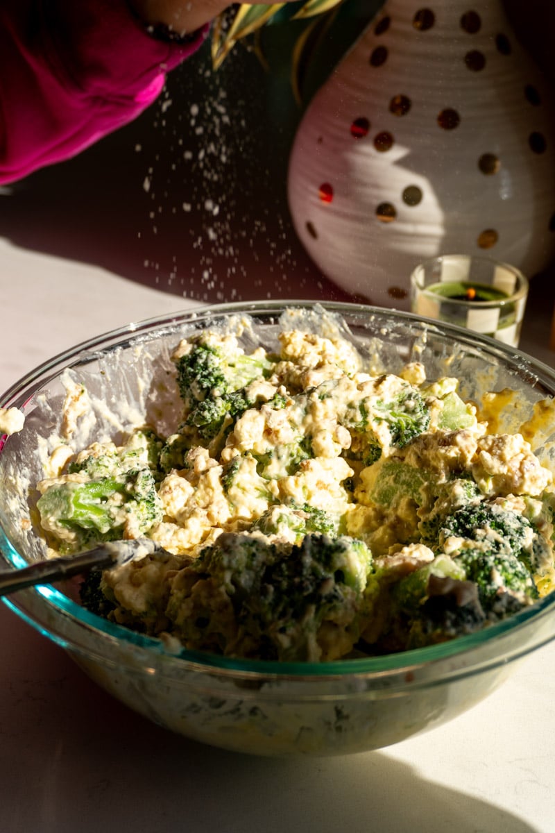 mackenzie smith sprinkling some salt into a glass bowl filled with creamy cheesy broccoli.